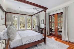 A four-poster bed with a wooden headboard, a desk with a chair, and a view of the outdoors through large windows.