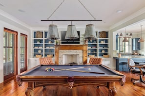 A pool table with cues, surrounded by leather chairs, in a room with a brick fireplace, shelves with decorative items, and hanging metal lamps.