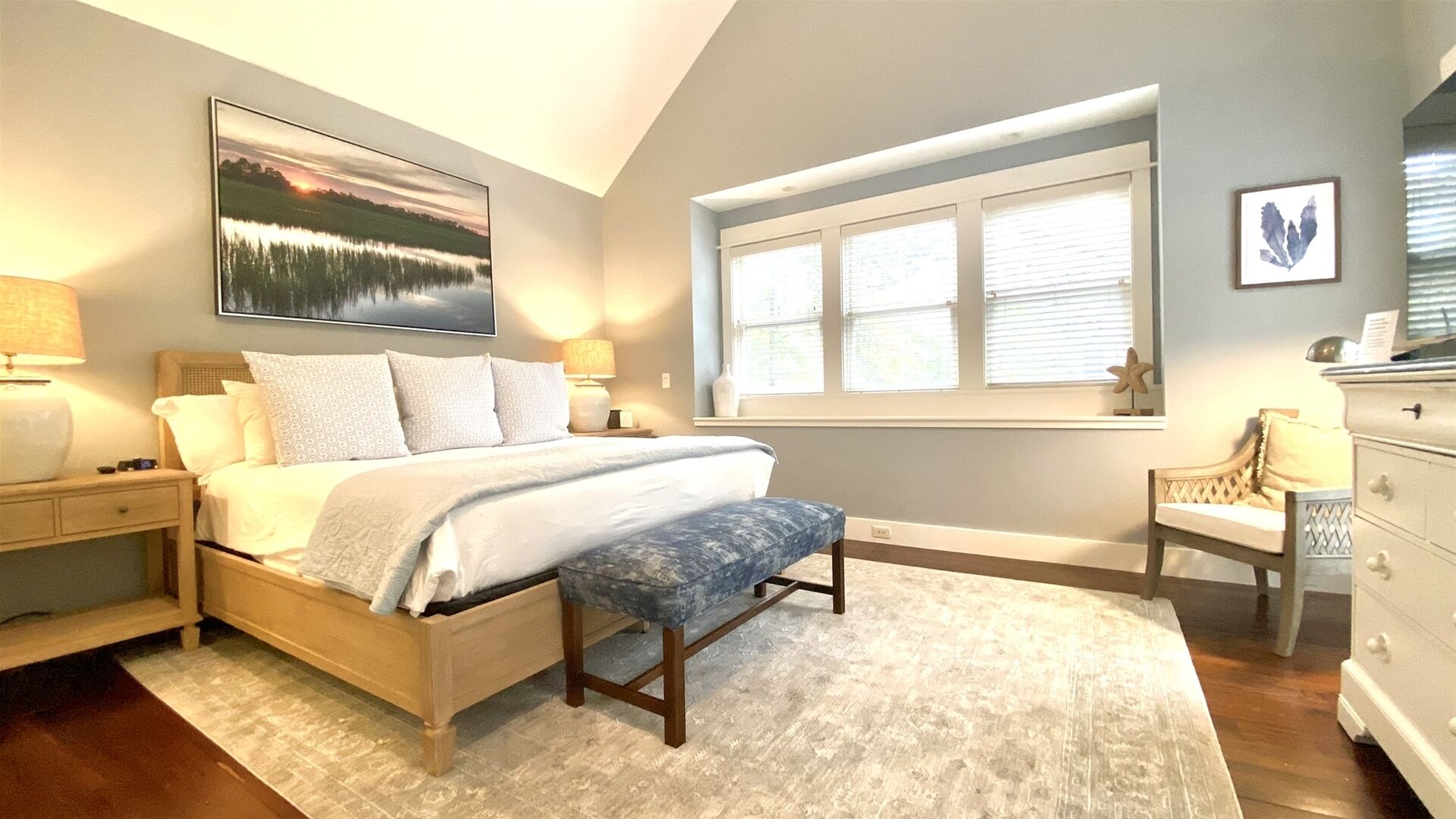 A bedroom with a bed, bedside tables, a bench, a dresser, a window with blinds, and a painting on the wall.
