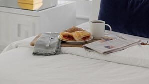 En snyggt bäddad säng med en kudde, ett nattduksbord med en lampa, en tallrik med mat, en kopp och en tidning.