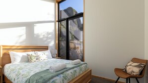 A bedroom with a large window, a wooden bed, a chair, and a nightstand. - Trails End (Punakaiki)