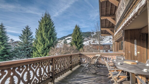 Um deck de madeira com guarda-corpo trabalhado, uma mesa com cadeiras e vista para colinas e árvores cobertas de neve.