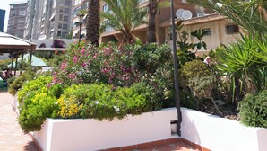 Un edificio de gran altura con varios balcones y un jardín bien cuidado con palmeras y plantas con flores.
