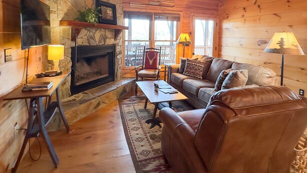 L’intérieur chaleureux d’un chalet avec une cheminée en pierre, des murs en bois, un canapé en cuir et un ventilateur de plafond.