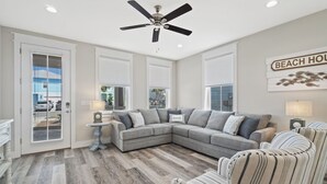 A modern living room with a gray sectional sofa, a ceiling fan, and a view of the outside through large windows.