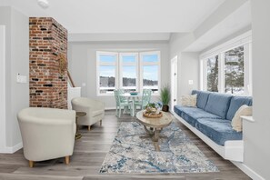 A spacious living room with a brick fireplace, a blue sofa, a dining area with a table and chairs, and large windows offering a view of the outdoors. - Exceptional 4BR Waterfront Home with Stunning Views, Waterfront Deck & Big Dock! (East Greenwich)