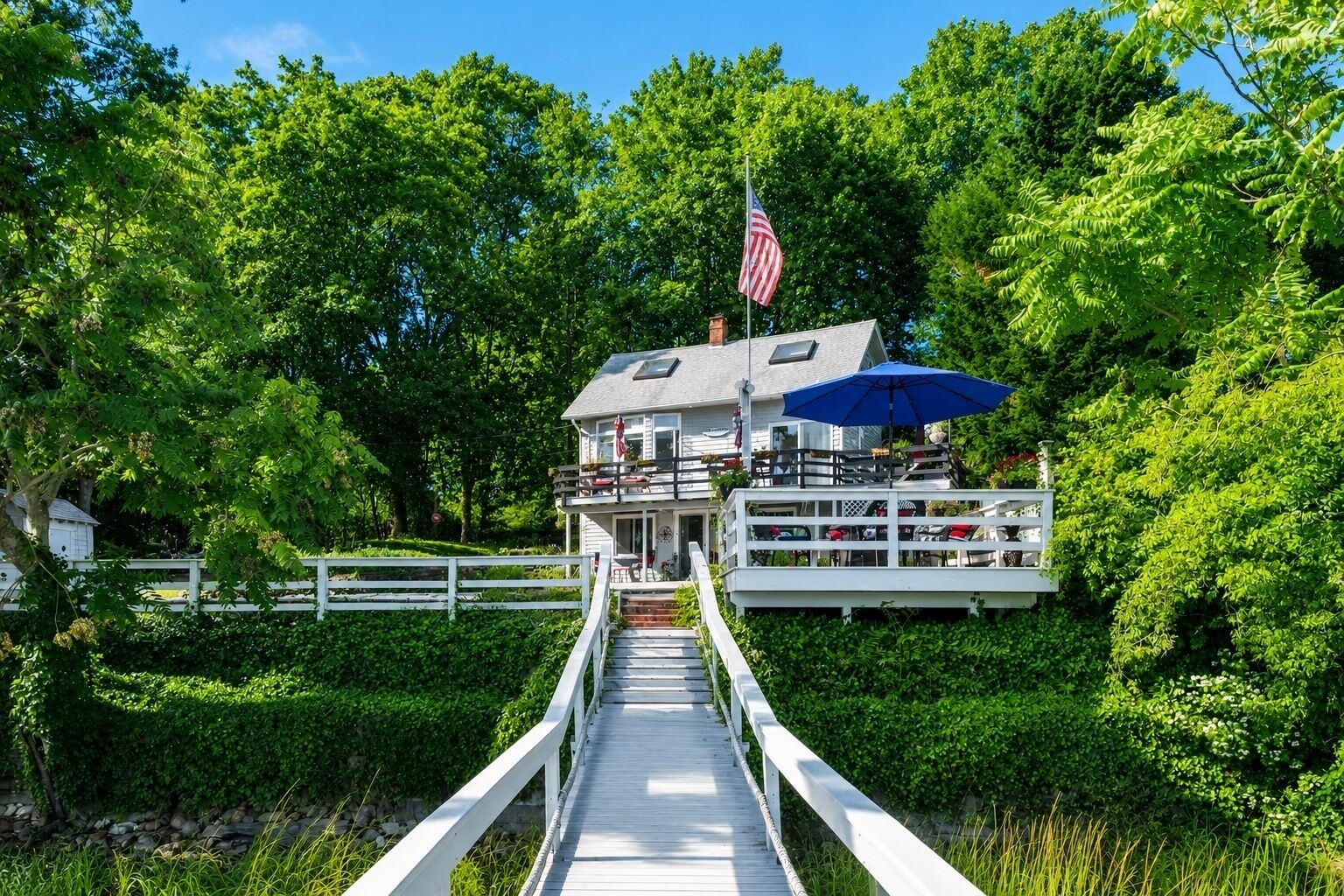 Een pand aan het water met een steiger die leidt naar een huis met een balkon, een Amerikaanse vlag en een blauwe parasol.
