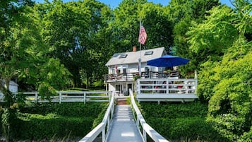 Een pand aan het water met een steiger die leidt naar een huis met een balkon, een Amerikaanse vlag en een blauwe parasol.