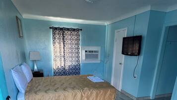 A bedroom with a bed, a nightstand, a lamp, a window with patterned curtains, an air conditioning unit, and a door.