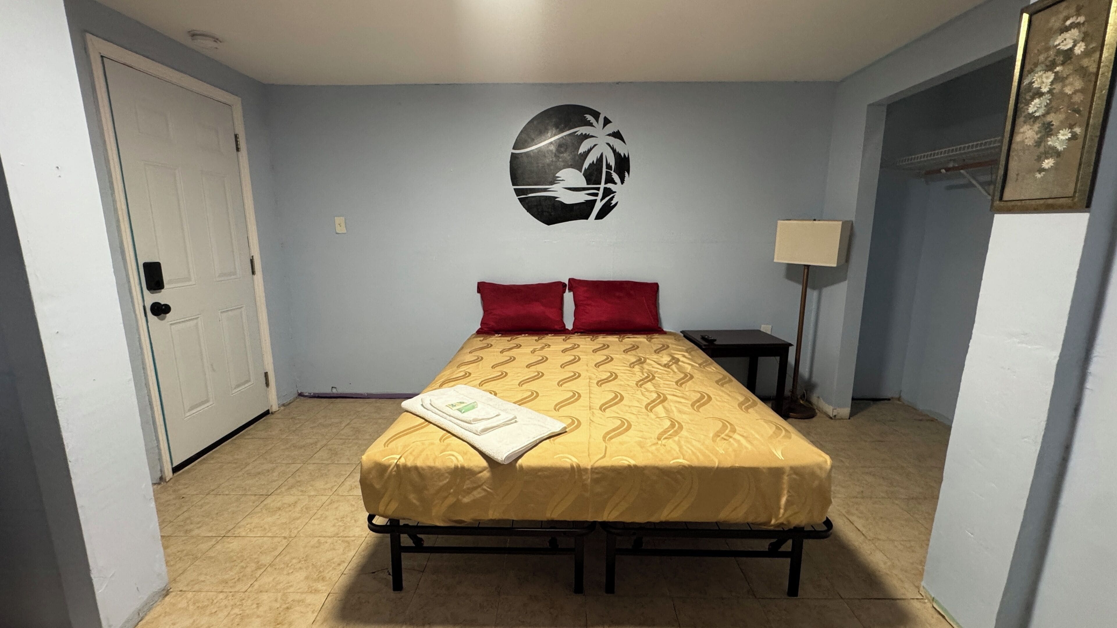 Un lit simple avec une couvre-lit à motifs, un oreiller rouge, une table de chevet, une lampe et un élément décoratif mural.
