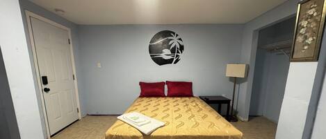 A single bed with a patterned bedspread, a red pillow, a bedside table, a lamp, and a wall decoration.