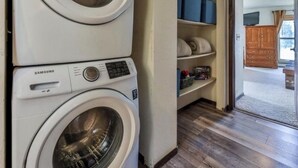 A laundry room with two stacked front-loading washing machines, a wooden floor, and a view into a living area with a fireplace. - Adorable 2-bedroom condo in tranquil Winter Park Colorado. (Winter Park)