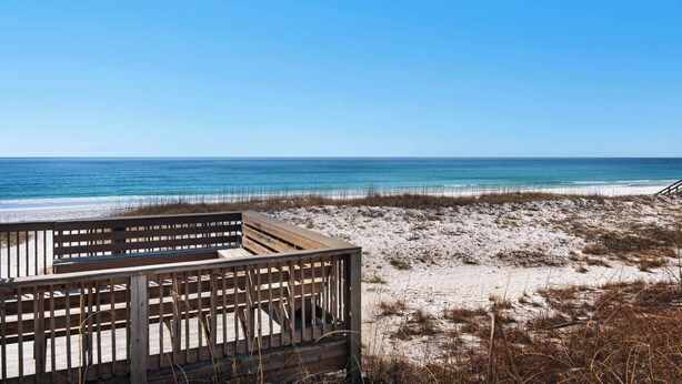 A beach view with a wooden deck and a sandy area with sparse vegetation. - Steps to the Beach Pool  Courts  Family Fun (Destin)