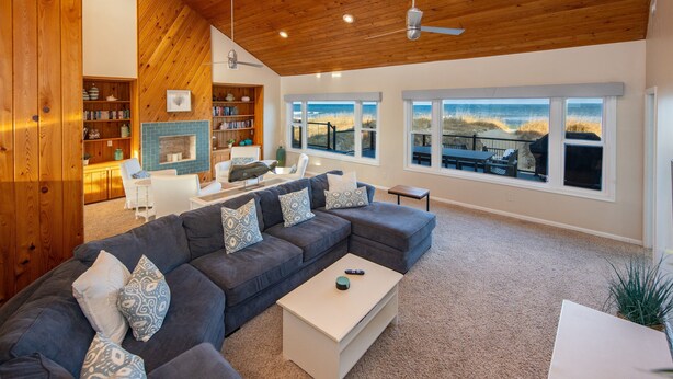 A spacious living room with a large sectional sofa, a fireplace, and large windows offering a view of the beach. - The Good Stuff - Oceanfront charmer with direct beach access (Virginia Beach)