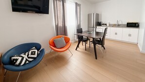 Una sala de estar moderna con televisión de pantalla plana, una mesa de comedor con sillas, una zona de cocina con refrigerador y microondas, y un sillón azul con un cojín estampado.