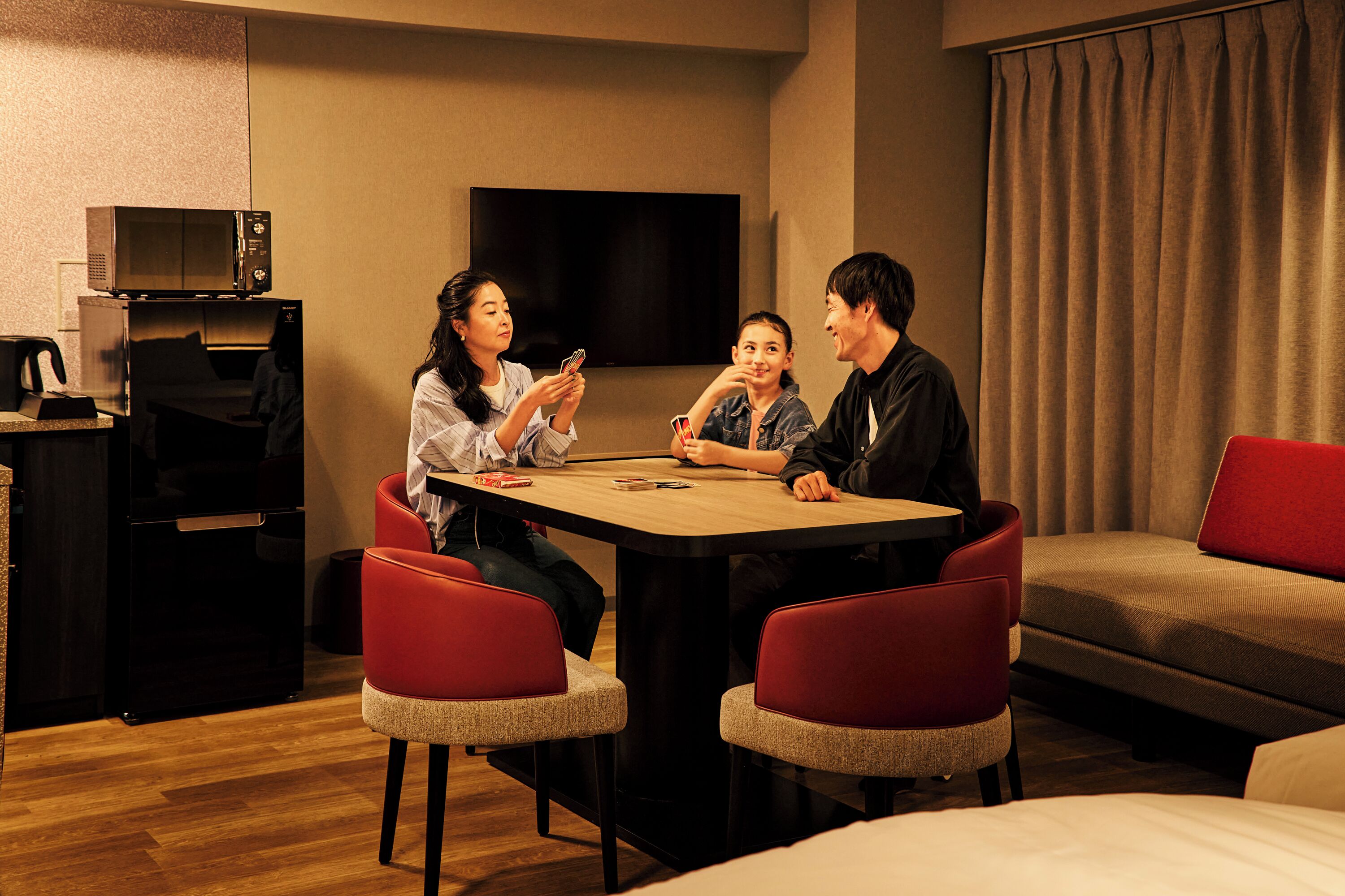 A hotel room with a wooden table, red chairs, a flat-screen TV, a sofa, and a bed.