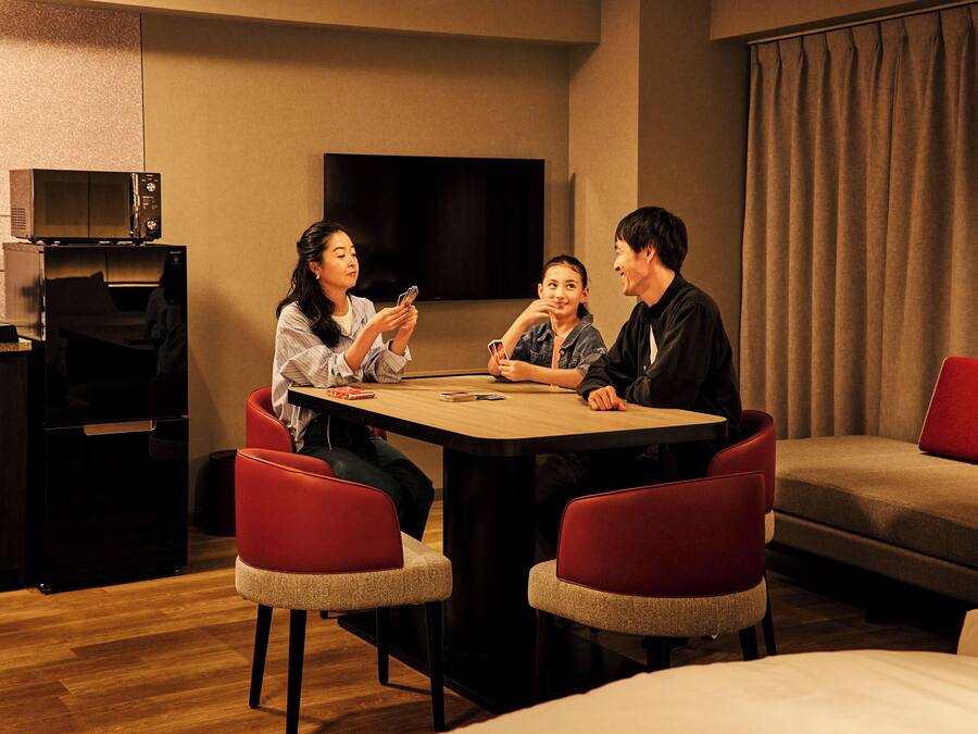 A hotel room with a wooden table, red chairs, a flat-screen TV, a sofa, and a bed.