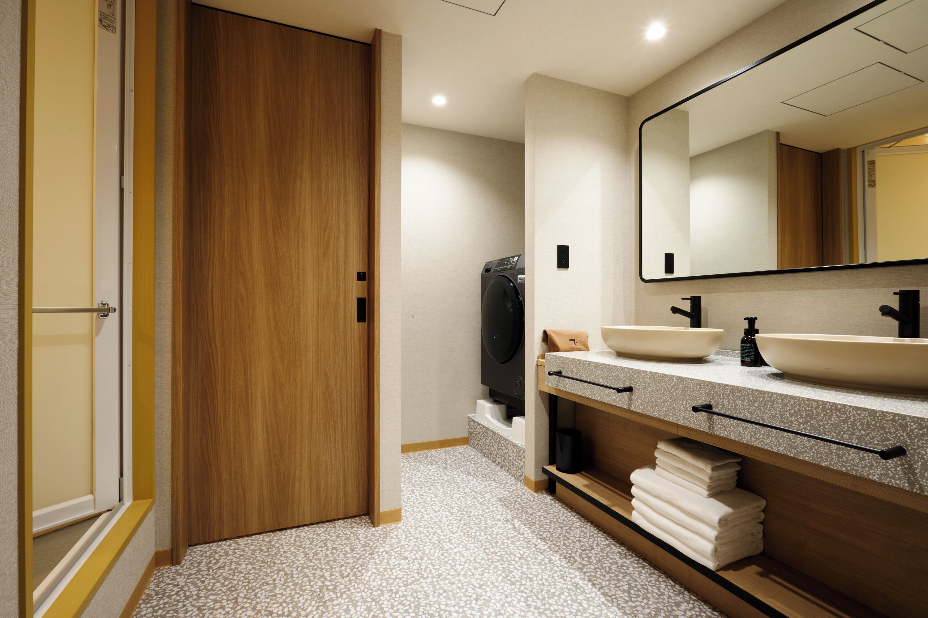 A modern bathroom with a wooden door, a large mirror, two sinks, and a towel rack.