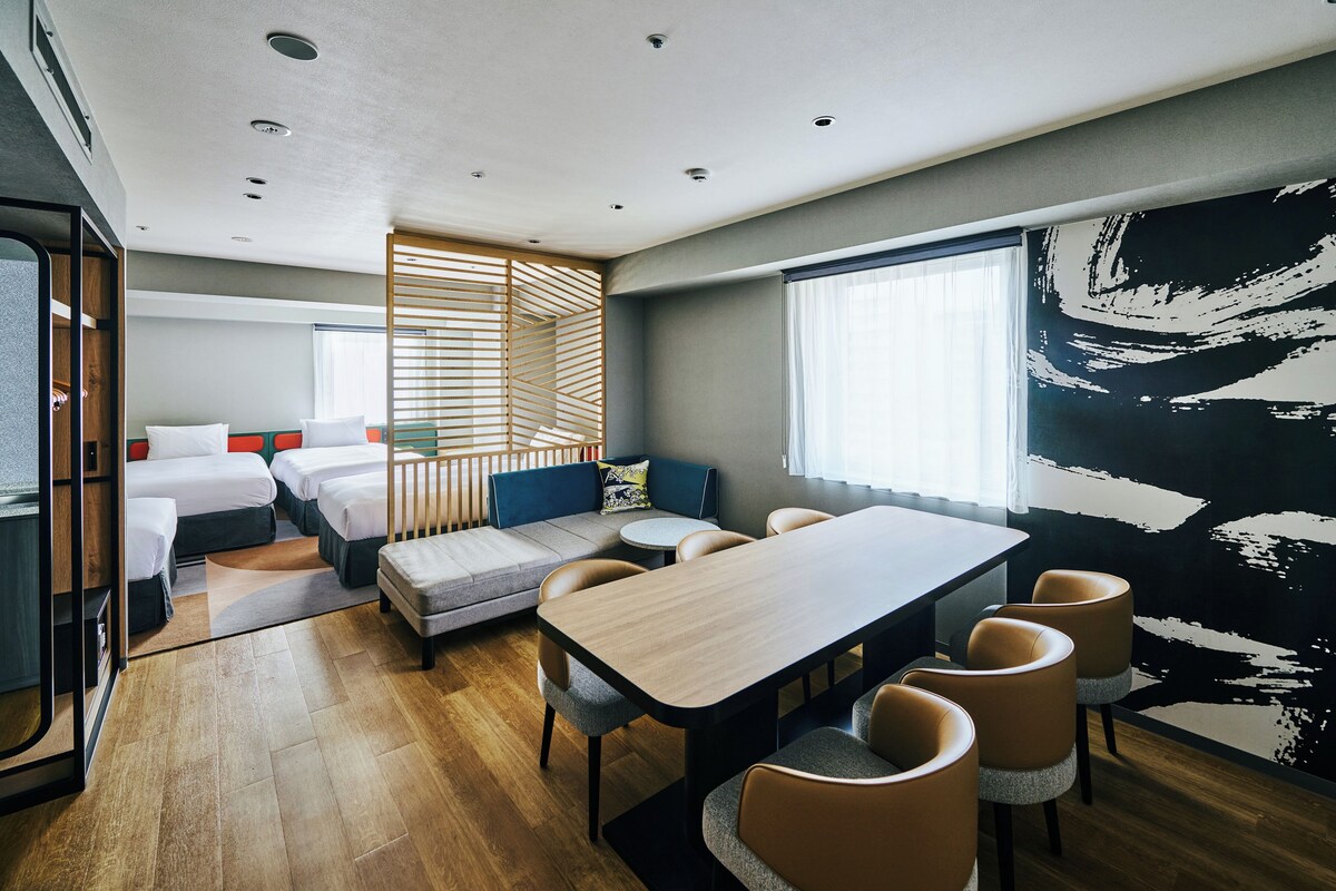 A modern hotel room with a large bed, a sofa, a dining table, and a large window.