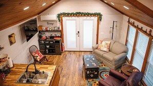 A cozy attic room with a wooden ceiling, a kitchenette, a sofa, and a window with blinds. - Marley: Cozy tiny cabin on Christmas Tree Farm in South Georgia (Cairo)