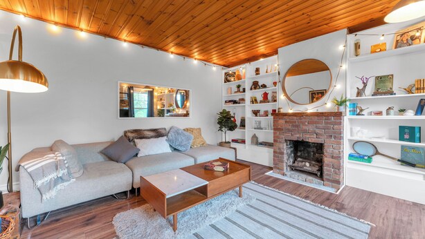 Een knusse woonkamer met een houten plafond, een schouw van baksteen, een bank en een boekenkast.