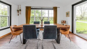 Een eetruimte met een houten tafel, vier stoelen en uitzicht op groen door grote ramen.