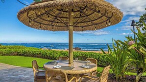 A round table with wicker chairs and a thatched umbrella, overlooking a beach with the ocean in the background. - Waipouli Beach Oceanfront Resort (Kapaa)