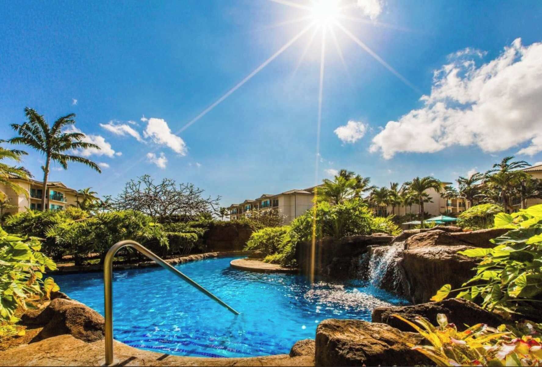 A pool area with a waterfall, surrounded by lush greenery and palm trees.