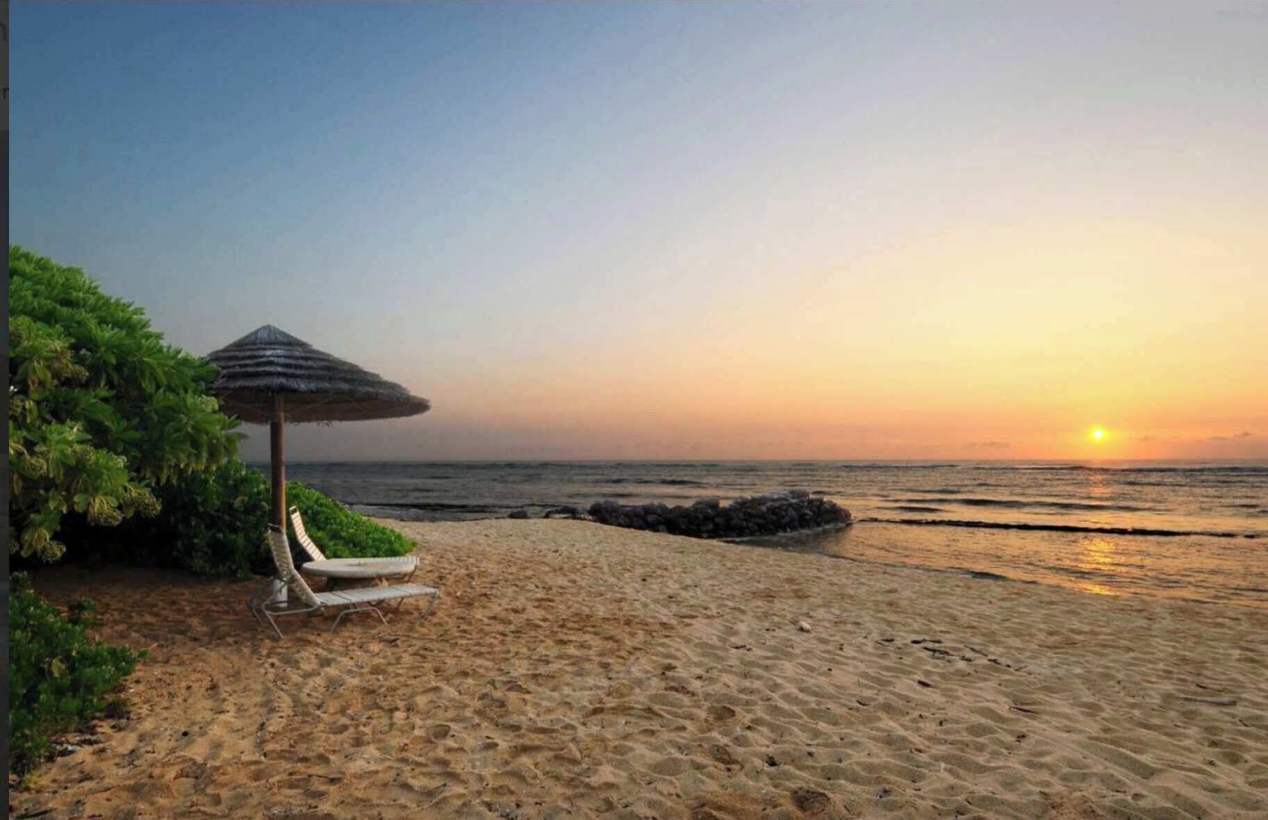 A beach with a thatched umbrella and lounge chairs, a rocky breakwater, and the ocean at sunset.