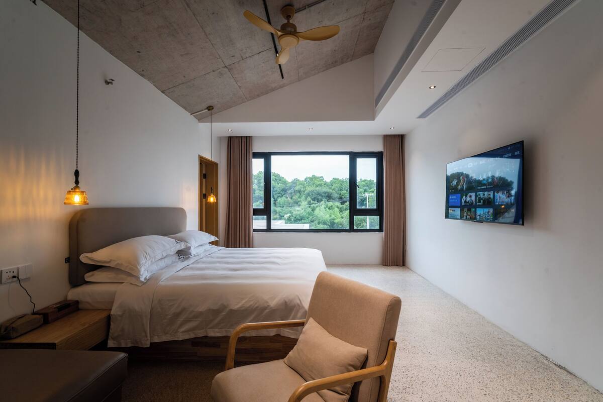 A hotel room with a bed, a chair, a TV, and a window with a view of greenery.