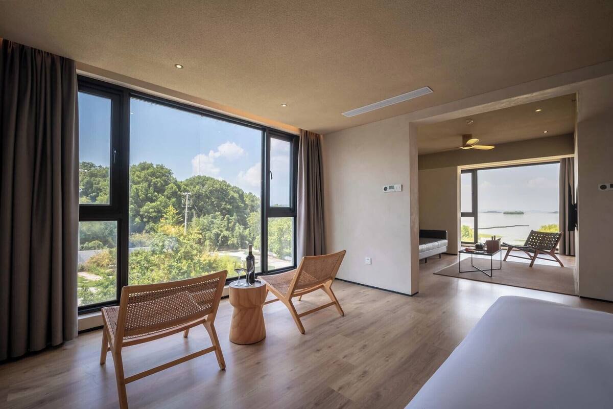 A spacious room with large windows, a bed, two wicker chairs, a small round table, and a view of a forested area.