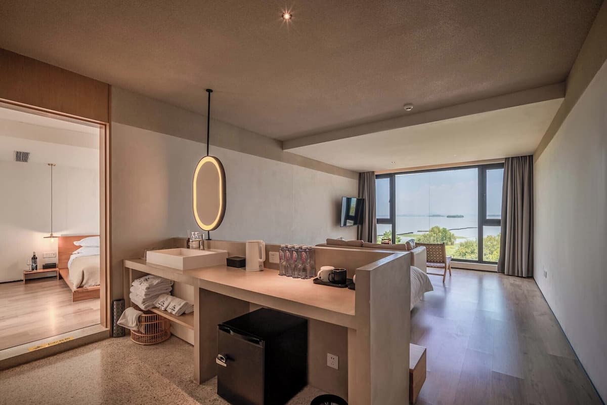 A modern hotel room with a large window, a built-in desk, a mirror, and a sofa.