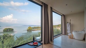A modern room with a large window overlooking a lake, a bathtub, and a table with a bottle and glasses.