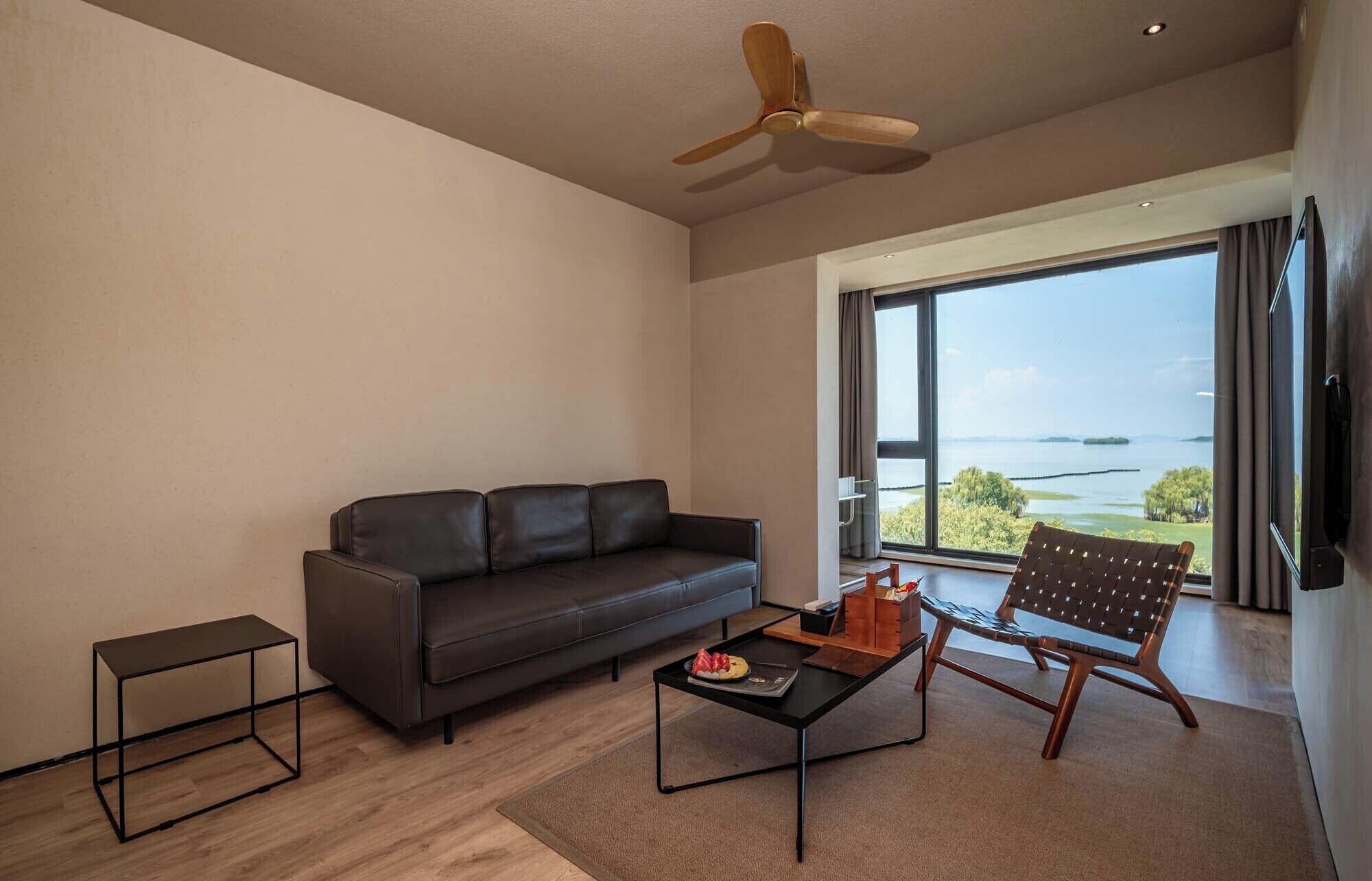 A modern living room with a leather sofa, a coffee table with a plate of strawberries, a wooden chair, and a view of a body of water through large windows.