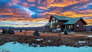 Une cabane en bois dotée d’une grande façade vitrée et d’un toit en métal, entourée d’un paysage enneigé et de petits arbres, le tout se détachant sur un ciel flamboyant au coucher du soleil.
