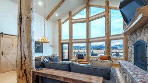 Un salon spacieux avec un grand canapé d’angle, une cheminée en pierre et une vue sur les montagnes grâce à de grandes fenêtres.