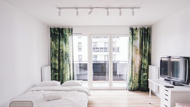 A modern hotel room with a bed, a desk with a TV, and a view of buildings through large windows. - Lubicka 69 Urban Retreat Apartment (Toruń)