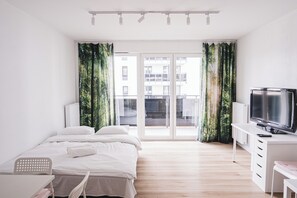 A modern hotel room with a bed, a desk with a TV, and a view of buildings through large windows. - Lubicka 69 Urban Retreat Apartment (Toruń)