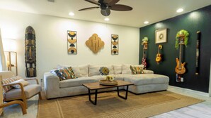Ein Wohnzimmer mit einer Couch, einem Couchtisch und Sesseln. An den Wänden h ängen Gitarren und es gibt einen Deckenventilator.