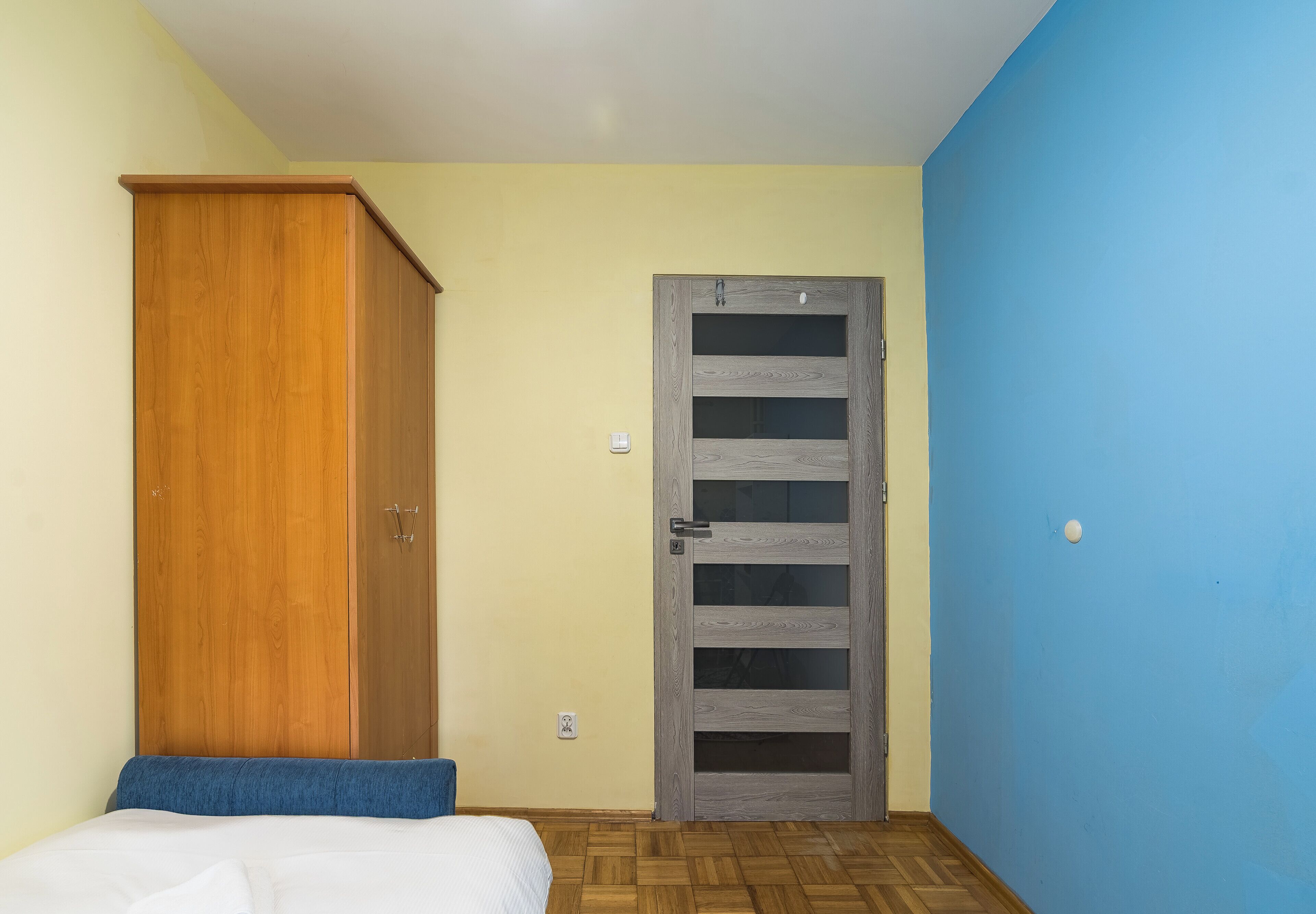 Ein Schlafzimmer mit einem Holzschrank, einem Bett mit blauer Kopfteil und einer grauen Tür mit Glasschiebeelementen.