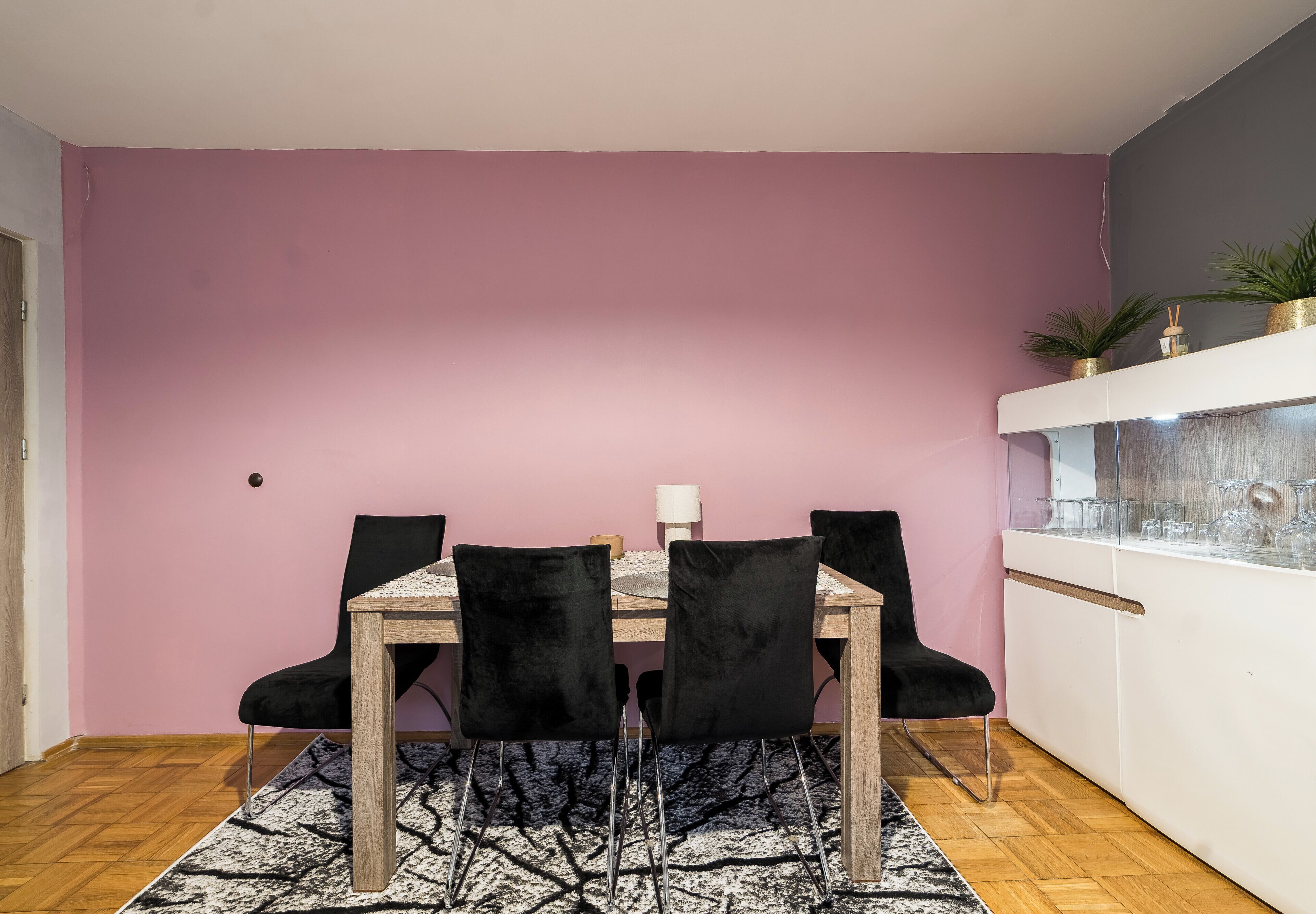 Uma área de refeições com uma mesa de madeira e quatro cadeiras pretas, uma parede rosa e um armário branco com copos.