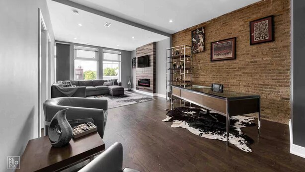 A modern living room with a grey sofa, a glass coffee table, and a leather chair. There is a fireplace, a TV mounted on the wall, and a wooden coffee table with a cowhide rug.