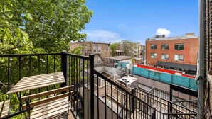 A balcony with a wooden bench, a metal railing, and a view of a red brick building and a blue construction site in the background.