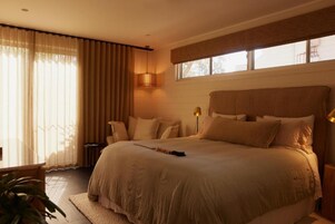 Uma cama grande com cabeceira, mesa de cabeceira, luminária e janela com cortinas.