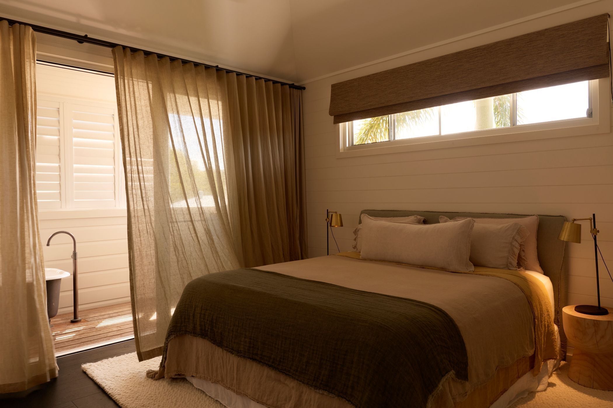 A bedroom with a bed, curtains, a window with blinds, a bedside lamp, and a small table.