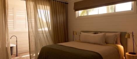 A bedroom with a bed, curtains, a window with blinds, a bedside lamp, and a small table.