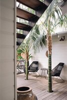 Una terraza de madera con dos sillas negras y una mesita pequeña, rodeada de palmeras y una pared blanca.