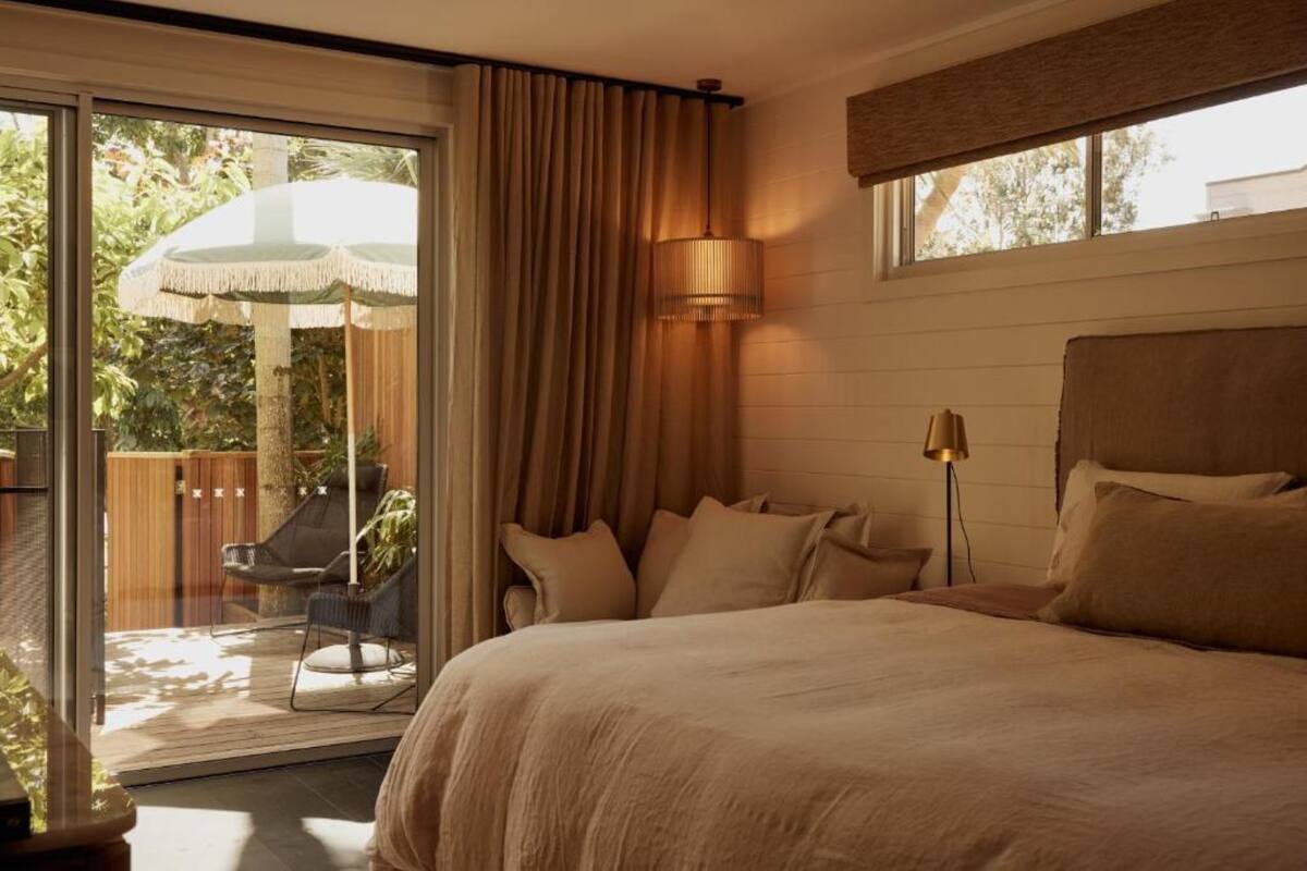 Een slaapkamer met een groot bed, nachtkastje en lamp. Er is een glazen schuifdeur naar een buitenruimte met een stoel en parasol.