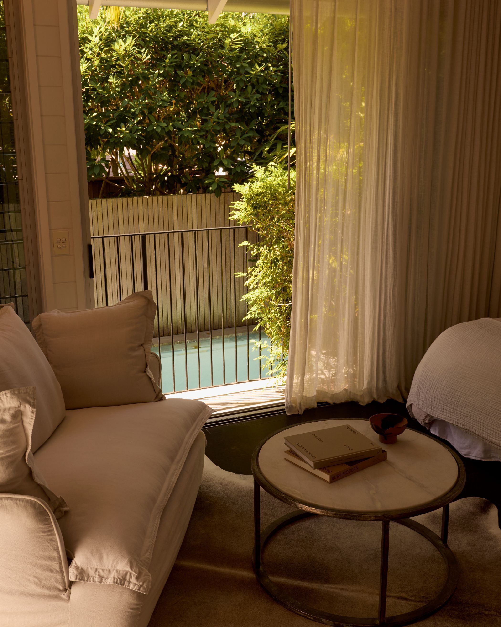 A room with a sofa, a round table with a book, and a view of a pool and greenery outside.