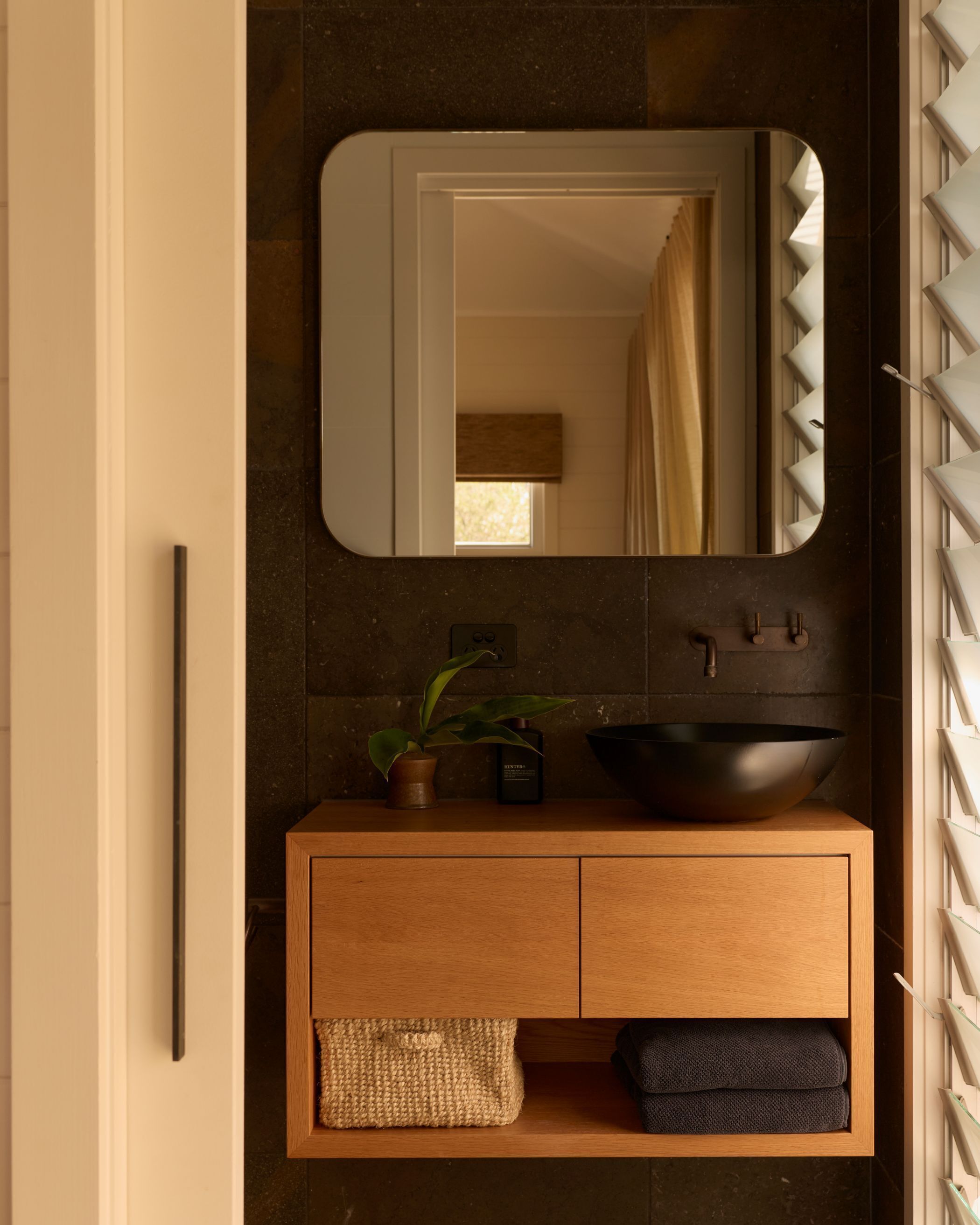 Een badkamer met een houten wastafel, een zwarte wasbak, een spiegel, een potplant en een handdoekrek.